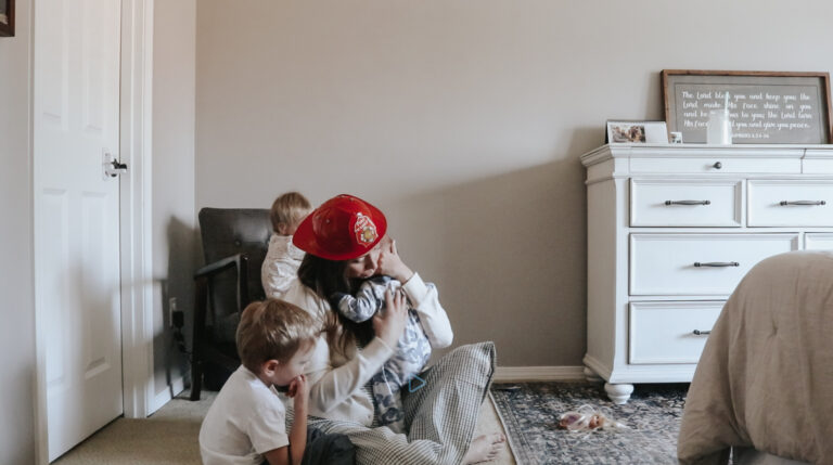 Mother wears a toy firehat and plays with her small children. She is hugging her littlest child.